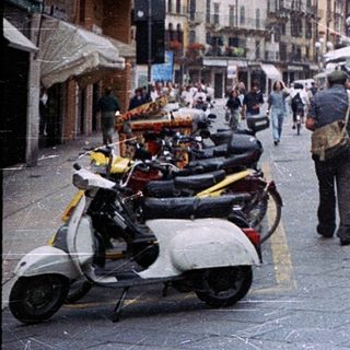 Historic European city street with pedestrians, scooters, and market stalls.