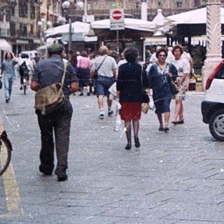 Historic European city street with pedestrians, scooters, and market stalls.