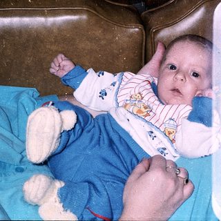 Baby in bathtub with teddy and hands visible.