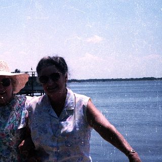 Two women smiling by water, wearing summer attire.