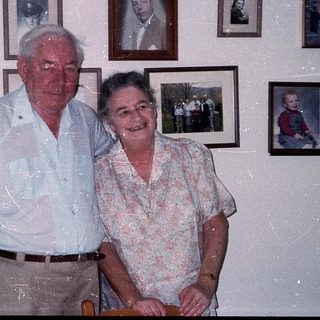 Two elderly couple posing together in a room with framed photos.