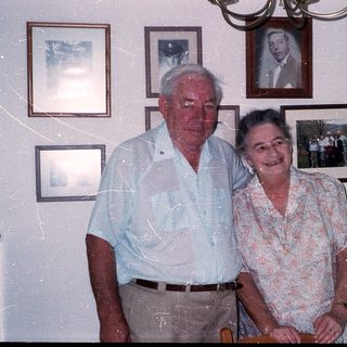 Two elderly couple posing together in a room with framed photos.