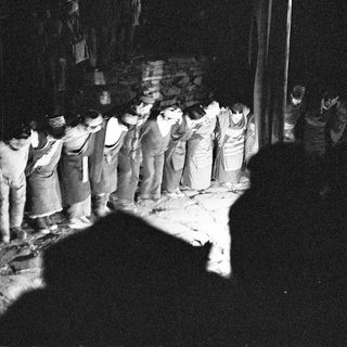 Group of people in traditional attire bowing during nighttime ceremony.