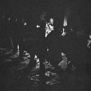 Dark outdoor gathering with people around a central figure playing guitar.