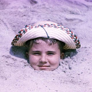 Child peeking out from sand with straw hat.