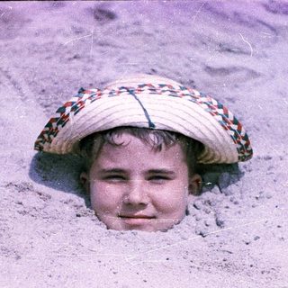 Child peeking out from sand with straw hat.