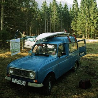 Classic blue van with surfboard, parked in forested area.
