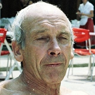 Shirtless older man sitting outdoors near umbrellas.
