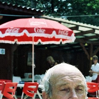 Shirtless older man sitting outdoors near umbrellas.
