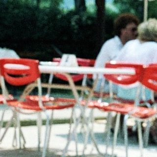 Shirtless older man sitting outdoors near umbrellas.
