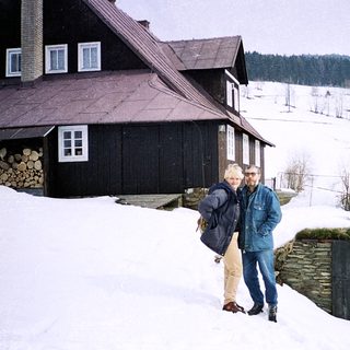 Two people standing in snowy rural landscape near wooden house.