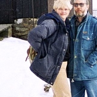 Two people standing in snowy rural landscape near wooden house.