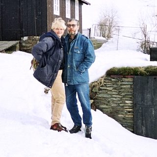 Two people standing in snowy rural landscape near wooden house.