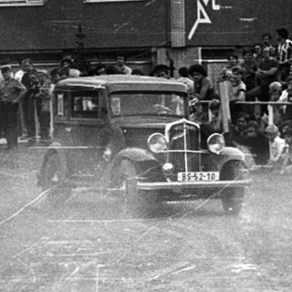 Historic car race on dirt track with spectators.
