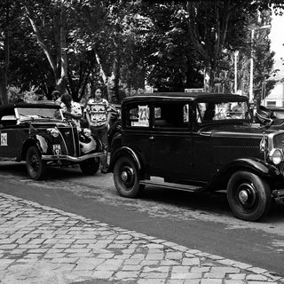 Vintage cars parked on cobblestone street with people.