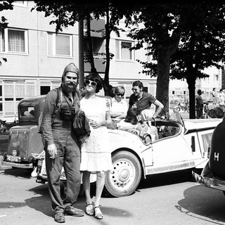 Vintage photo: man in military uniform with woman in 1940s attire near vintage cars.