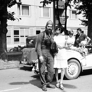 Vintage photo: man in military uniform with woman in 1940s attire near vintage cars.