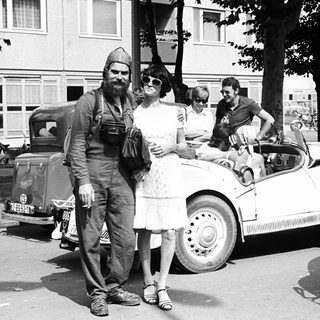 Vintage photo: man in military uniform with woman in 1940s attire near vintage cars.