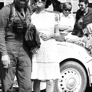 Vintage photo: man in military uniform with woman in 1940s attire near vintage cars.