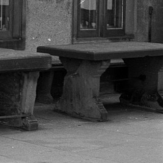 Old woman sitting alone on weathered bench outside aged building.
