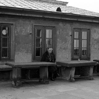Old woman sitting alone on weathered bench outside aged building.