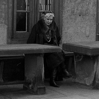 Old woman sitting alone on weathered bench outside aged building.