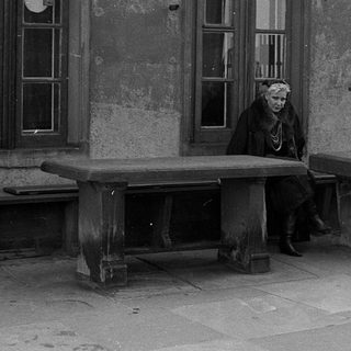 Old woman sitting alone on weathered bench outside aged building.