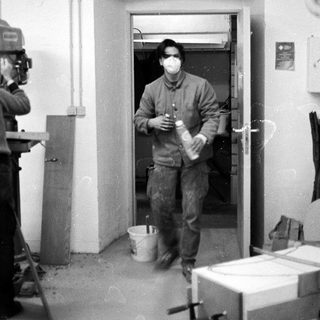 Man in protective gear holding container, exiting through doorway under filming.