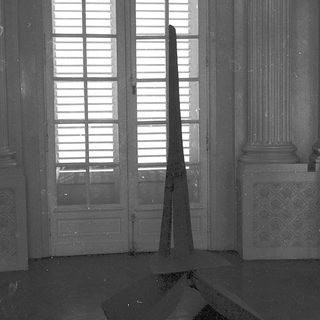 Empty room with tall pyramid sculpture and open shuttered windows.