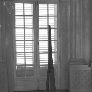Empty room with tall pyramid sculpture and open shuttered windows.