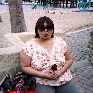 Woman sitting outdoors near beach, wearing sunglasses and floral blouse.