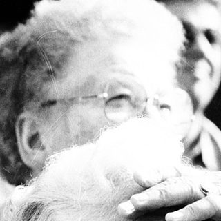 Elderly man adjusting sunglasses indoors with crowd.
