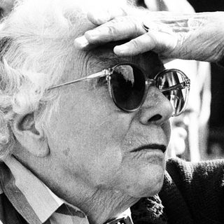 Elderly man adjusting sunglasses indoors with crowd.