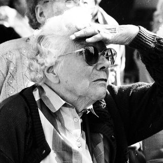 Elderly man adjusting sunglasses indoors with crowd.