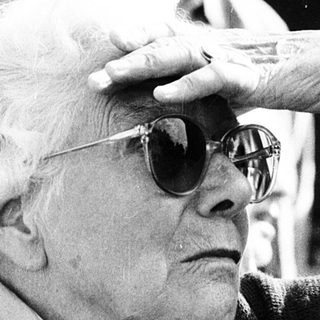 Elderly man adjusting sunglasses indoors with crowd.