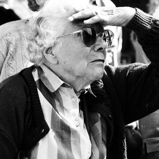 Elderly man adjusting sunglasses indoors with crowd.