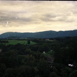 Rural landscape with hills, forest, and farmland under cloudy sky.