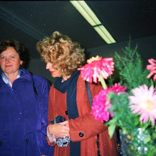 Two women standing indoors near floral arrangement.