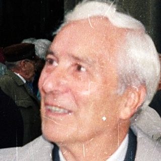 Elderly man in coat holding umbrella at outdoor event.