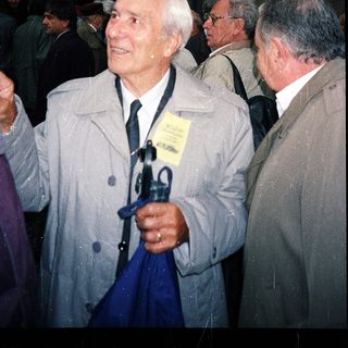 Elderly man in coat holding umbrella at outdoor event.