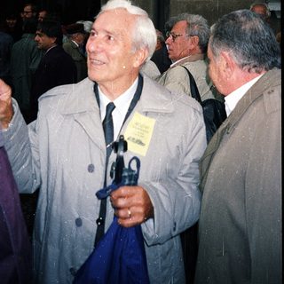 Elderly man in coat holding umbrella at outdoor event.