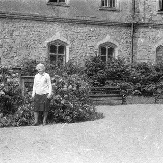 Old woman standing outdoors near historic stone building.