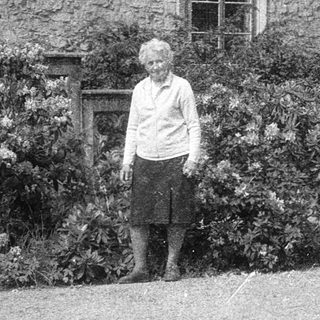 Old woman standing outdoors near historic stone building.