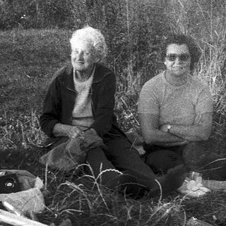 Two elderly people sitting outdoors with dog, surrounded by household items and firewood.