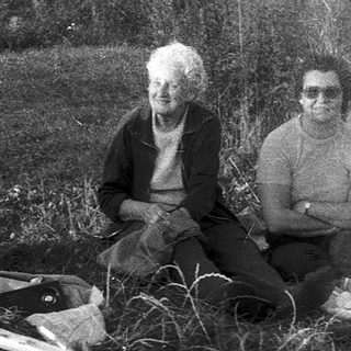 Two elderly people sitting outdoors with dog, surrounded by household items and firewood.