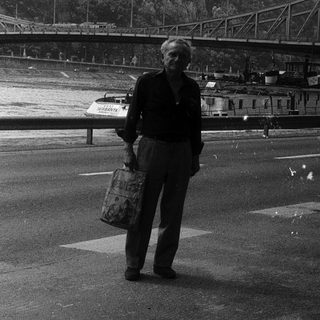 Man standing by waterfront with bridge and hilltop monument.