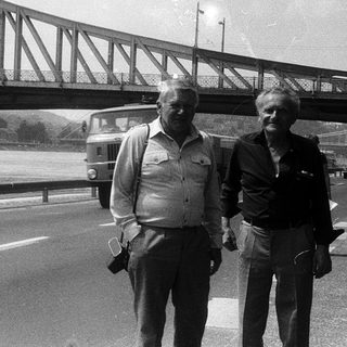 Two men walking near a suspension bridge.