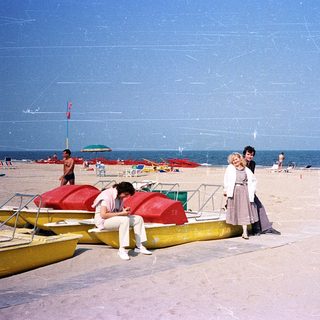 Beach scene with people sunbathing, boats, and umbrellas.