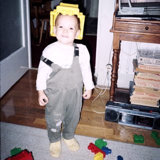 Child wearing yellow Lego helmet and overalls indoors.