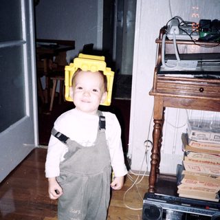Child wearing yellow Lego helmet and overalls indoors.